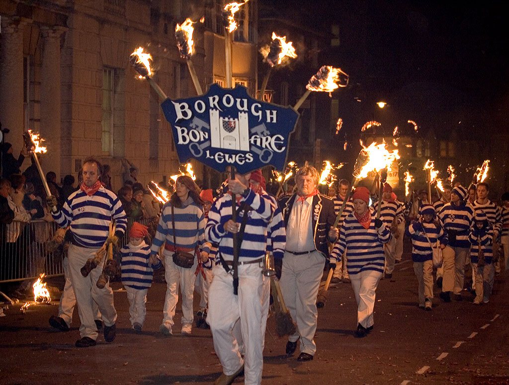 Members of the Lewes Borough Bonfire Society in striped jerseys march with flaming torches behind their society banner